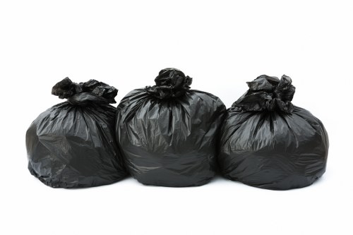 Three large black garbage bags made of plastic are positioned in a row on a plain white background, with the bags tightly knotted at the top. The bags appear to be filled with waste or rubbish, showing slight bulges and irregular shapes indicating their contents. The surface of the plastic bags has a matte finish with some creases and wrinkles visible. The bags are situated in a neutral environment without any additional objects or surroundings, emphasizing the typical appearance of waste ready for disposal or collection. This visual aligns with rubbish collection services provided by companies such as Business Waste Removal Chiswick, and the setting suggests an indoor or outdoor setting suitable for waste handling. The composition focuses solely on the bags, conveying the concept of waste management visually suitable for a professional rubbish removal context in the Chiswick area.
