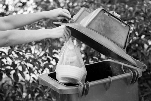 A person’s hands are seen lifting the lid of a grey outdoor rubbish bin situated on a paved driveway, revealing the interior. Inside the bin, there is a clear plastic bag filled with household waste, including white paper or cardboard containers. The bin has a black interior and a slightly textured finish on its exterior, which appears to be made from durable plastic. In the background, blurred greenery suggests a garden or outdoor area typical of a residential property. The image is in black and white, highlighting the contrast between the bin’s textured surface and the smooth plastic waste bag. This scene is representative of waste collection or rubbish disposal activities commonly performed by rubbish removal services, such as those offered by Business Waste Removal Chiswick, illustrating the process of emptying outdoor waste bins in a suburban environment, potentially within the postcode area associated with Chiswick, London.