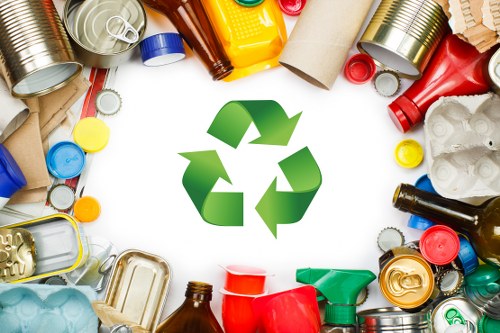 A circular arrangement of various recyclable waste items, including aluminum cans, plastic bottles, glass jars, cardboard tubes, and plastic containers, spread out on a white surface. The items display diverse textures and colours, such as shiny metallic surfaces, translucent plastics, and rough cardboard, with some cans and bottles positioned upright and others lying on their sides. In the center of the image, a prominent green recycling symbol made of three arrows pointing clockwise emphasizes the concept of waste recycling. The scene suggests a focus on waste separation and environmental responsibility, relevant to rubbish removal services in Chiswick, where Business Waste Removal Chiswick operates. The clear lighting and neat organization highlight the importance of managing different types of waste for sustainable disposal and recycling efforts in a local context.
