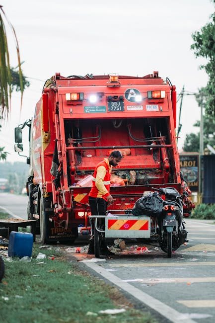 A male waste collection worker wearing a high-visibility yellow and red vest is operating a large red garbage truck on a street, positioned in front of the truck's rear collection hatch. The worker ap