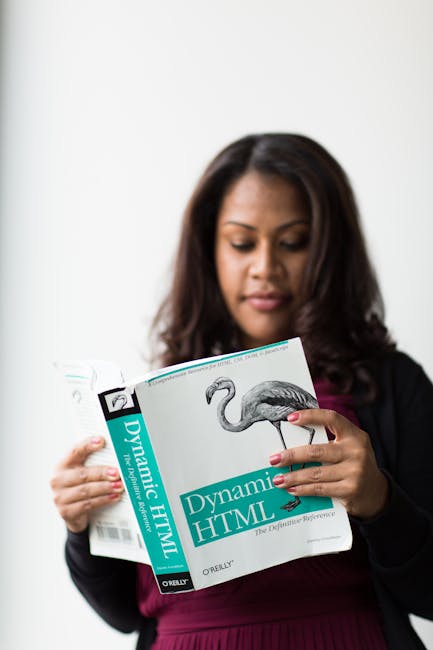 A woman with medium-length dark brown hair, dressed in a black blazer over a dark red top, is holding a book titled 'Dynamic HTML' by O'Reilly. She is looking down attentively at the book, which featu